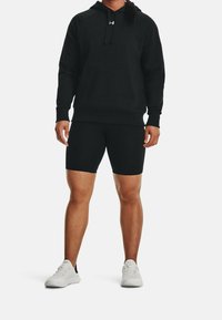 Sweat à capuche noir avec poche avant ; associé à un short noir. Les deux sont en tissu lisse. Des baskets blanches avec des accents en mesh complètent la tenue.