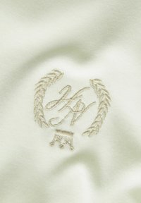 Broderet logo i cremefarvet tråd på blødt, lyst beige stof, med en laurbærkrans og initialerne "KH," med fine detaljer og glat tekstur.