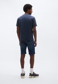 Granatowy T-shirt z krótkim rękawem, wykonany z bawełny, w połączeniu z ciemnymi szortami dżinsowymi, z widocznym przeszyciem na plecach i lekko zaokrąglonym dołem.