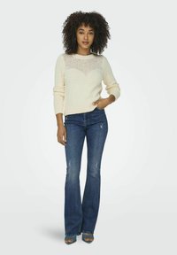 Pull en maille crème avec un motif texturé à l'encolure, assorti à un jean évasé sombre présentant des déchirures légères et un léger effet usé.