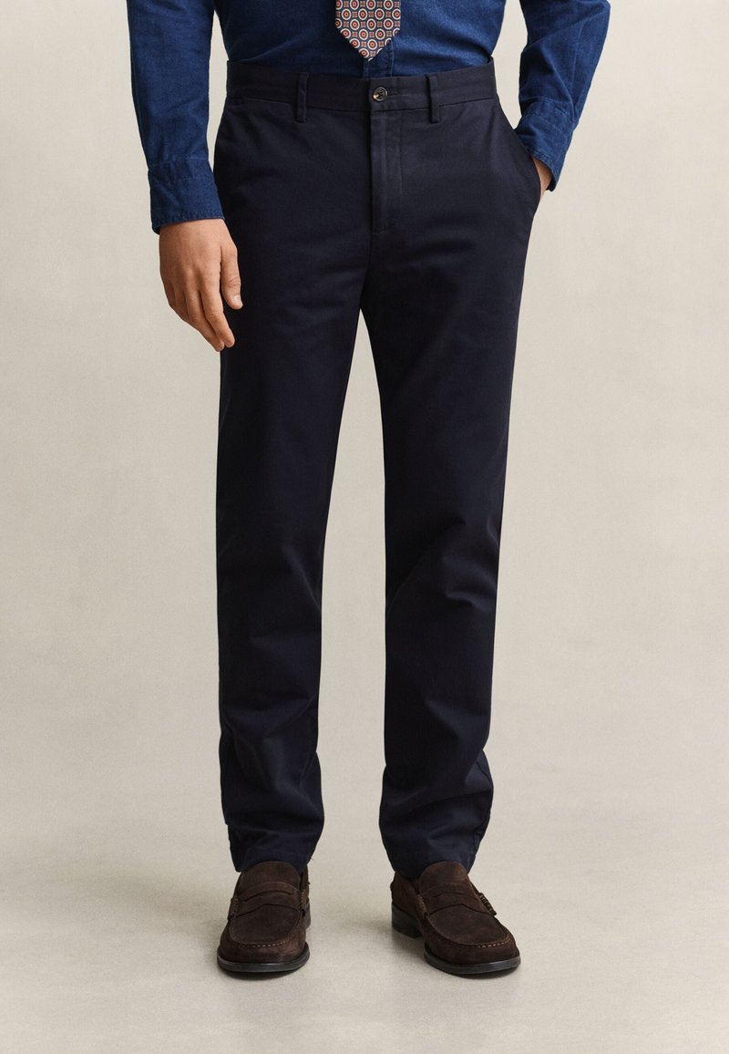 Homme portant un pantalon bleu marine foncé, des mocassins marron, une chemise bleue et une cravate à motif, debout avec une main dans la poche.
