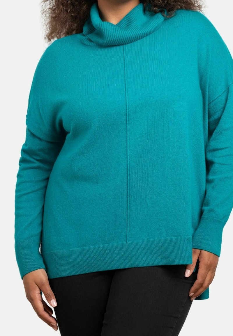Personne portant un pull en maille turquoise à manches longues avec un col bénitier et un pantalon noir, visible des épaules aux hanches sur fond blanc.