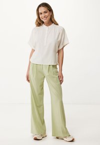 Blusa bianca con colletto e maniche corte, abbinata a pantaloni larghi verde chiaro; vestibilità rilassata, texture liscia e design minimalista.