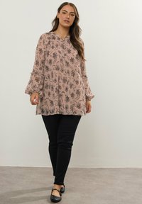 Blush pink tunika med sort blomsterprint, plisseret stof, rund halsudskæring og klokkeærmer, parret med tætsiddende sorte bukser og flade sko.