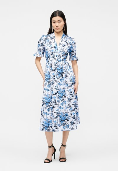 Γυναίκα που φοράει λευκό midi φόρεμα με μπλε floral σχέδιο, φουσκωτά κοντά μανίκια και μαύρα ψηλοτάκουνά πέδιλα με λουράκι στον αστράγαλο, στέκεται μπροστά σε απλό φόντο.