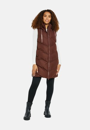 Braune Steppweste mit hohem Kragen, mit Reißverschluss, Vordertaschen und vertikalen Nähmustern; kombiniert mit schwarzen Leggings und Ankle Boots.