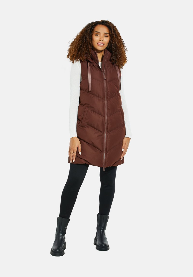 Braune Steppweste mit hohem Kragen, mit Reißverschluss, Vordertaschen und vertikalen Nähmustern; kombiniert mit schwarzen Leggings und Ankle Boots.