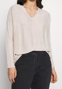 Jasnobeżowy sweter z długimi rękawami i fakturą prążkowaną, z dekoltem w serek. W zestawieniu z czarnymi jeansami z wysokim stanem. Sweter ma luźny krój i boczne rozcięcia.