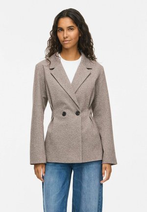 Frau mit langen lockigen Haaren, die einen taupefarbenen, zweireihigen Blazer über einem weißen Hemd und blauen Jeans trägt, steht vor einem schlichten weißen Hintergrund.