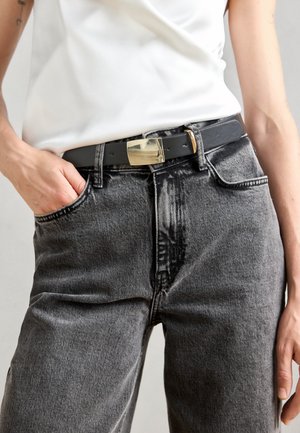 Juodas diržas su stačiakampiu auksiniu sagtimi, uždedamas ant aukštu juosmeniu dėvimų pilkų džinsų. Džinsai turi nublukusią tekstūrą ir priekines kišenes.