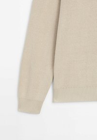 Manche et ourlet de pull tricoté beige avec poignets et ceinture côtelés sur fond blanc.