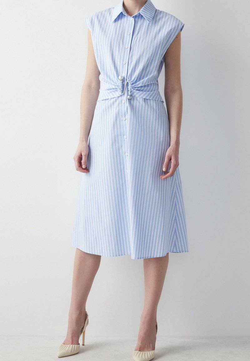 Femme portant une robe sans manches à rayures bleu clair et blanc avec une taille froncée et des chaussures beiges à talons hauts.