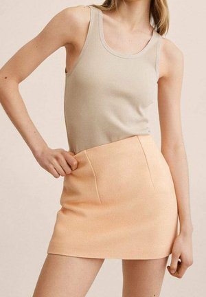 Femme portant un débardeur beige sans manches et une mini-jupe pêche taille haute, posant avec une main sur la hanche devant un fond neutre.