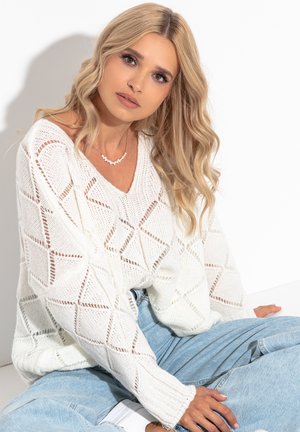 Biały sweter dzianinowy z diamentowym wzorem, głębokim dekoltem w kształcie V i luźnym krojem. Jasnoniebieskie dżinsy dopełniają swobodny wygląd.