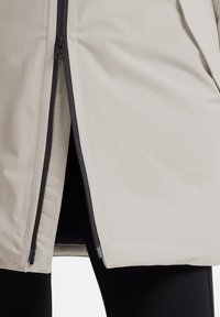 Giacca imbottita beige con una finitura liscia e opaca; presenta una zip nera lungo la cucitura frontale e tasche nascoste. Collant neri visibili sotto.