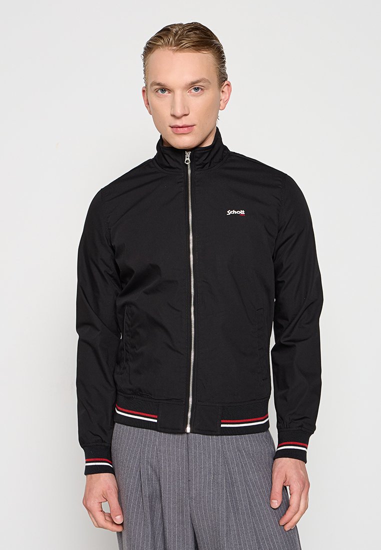 Veste noire zippée avec un col montant, présentant un petit logo sur la poitrine. Poignets et ourlet côtelés avec des rayures rouges et blanches. Texture lisse.
