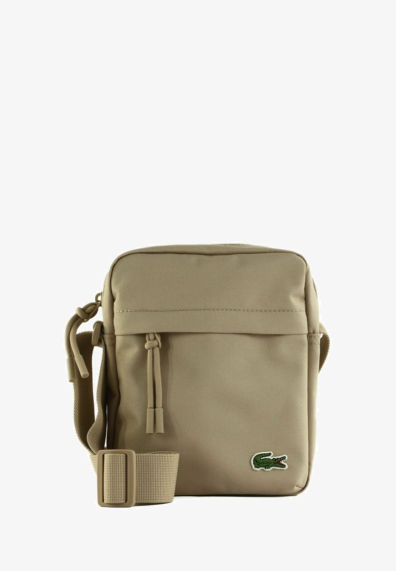 Borsa a tracolla beige con tracolla regolabile, tasca frontale con zip e piccolo logo di coccodrillo verde nell'angolo in basso a destra.
