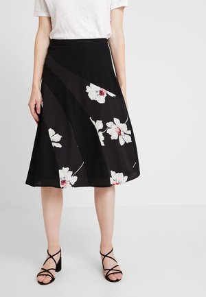 Femme portant une jupe noire trapèze avec de grands motifs floraux blancs et roses, associée à un T-shirt blanc et des talons noirs à lanières.