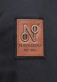 Etiqueta de cuero marrón con el logo en relieve "N" y acentos circulares, cosida sobre un fondo de tela oscura. El texto dice "NAVAHOO EST 2014."