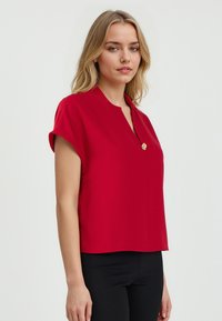 Blouse rouge à manches courtes avec un col mandarin, dotée d'un détail de bouton doré à l'avant. Tissu lisse avec une coupe ajustée.