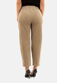 Femme debout portant un pantalon en velours côtelé beige et des talons hauts noirs, vue de dos sur un fond blanc.