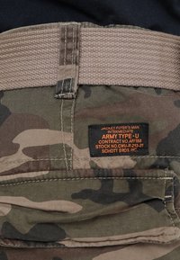 Pantalon cargo en tissu à motif camouflage avec une ceinture tissée beige et une étiquette noire détaillant les informations de fabrication.