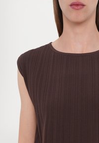 Femme portant un haut en tricot côtelé sans manches de couleur marron foncé, avec un col rond, visible des épaules jusqu’à juste en dessous du menton.