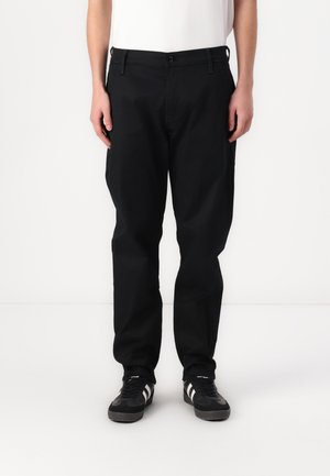 Persoon die losse zwarte broek en zwarte sneakers met witte strepen draagt, staand tegen een effen witte achtergrond.