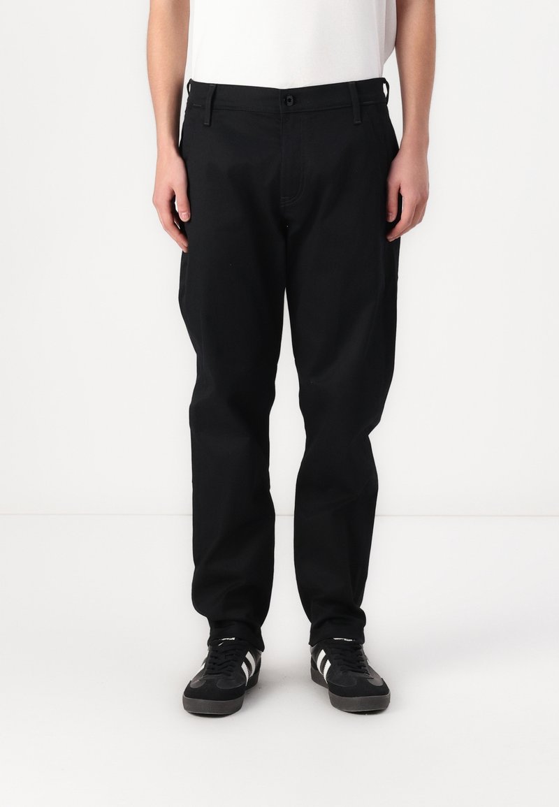 Personne portant un pantalon noir ample et des baskets noires avec des rayures blanches, debout devant un fond blanc uni.