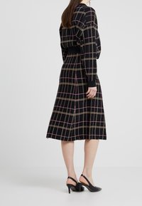 Vestido negro con patrón de cuadros multicolores, mangas largas y cintura elástica. Combinado con tacones slingback negros. Textura de tela suave.