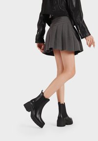 Botines negros con un acabado liso, paneles laterales elásticos y un tacón cuadrado. El conjunto incluye una falda gris con pliegues y una chaqueta de cuero.