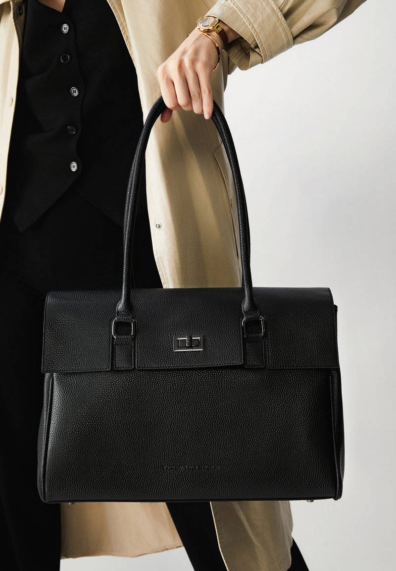 Mano sosteniendo un bolso de cuero negro texturizado con doble correa, vistiendo un abrigo beige y un reloj de pulsera dorado.