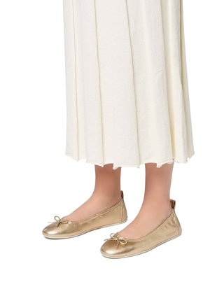 Ballerines métalliques dorées avec une texture lisse, ornées d'un nœud au niveau des orteils et d'une semelle blanche flexible, assorties à une jupe plissée blanche.