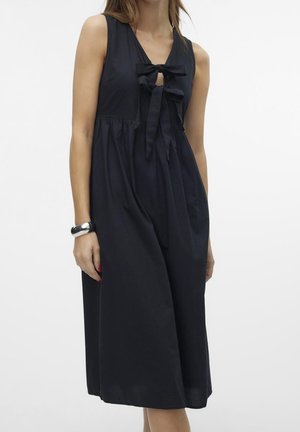 Vrouw draagt een mouwloze zwarte midi-jurk met twee strikken aan de voorkant en een geplooide taille, gecombineerd met een zilveren armband aan de linkerpols.