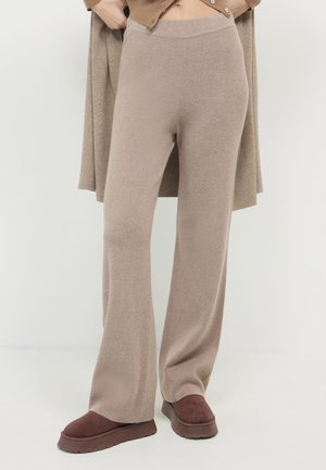 Pantalons tricotés beiges à jambes larges associés à des chaussures à plateforme marron foncé, portés par une personne se tenant devant un fond blanc.