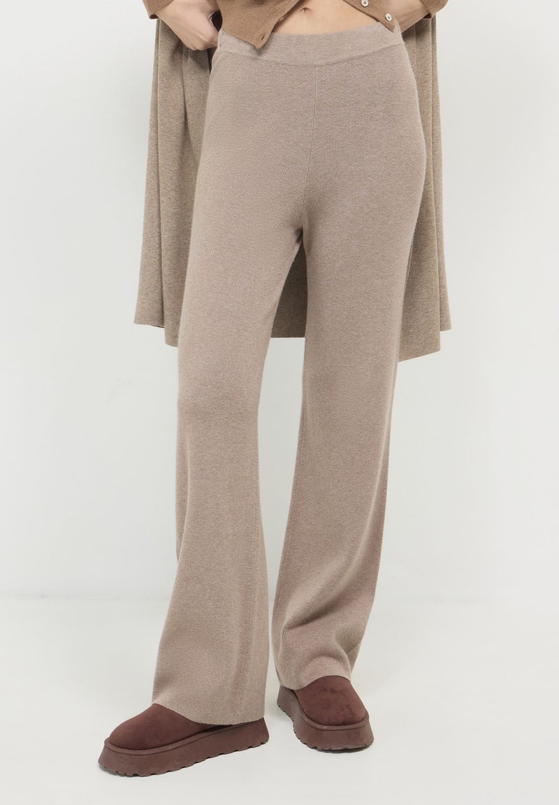 Beige weite Strickhosen kombiniert mit dunkelbraunen Plateau-Slippern, getragen von einer Person, die vor einem weißen Hintergrund steht.
