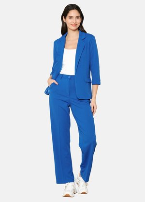 Ensemble composé d'un blazer bleu et de pantalons larges, fabriqué en tissu lisse. Il présente un revers à cran, une fermeture à bouton et des poches latérales. Porté avec un haut blanc.