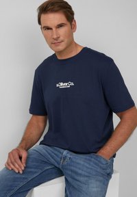 Donkerblauw T-shirt met korte mouwen van katoen, met een ronde hals en een witte logo-opdruk. Het model zit en draagt lichtblauwe jeans.