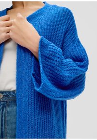 Modrý pletený kardigan s volným střihem, širokými rukávy a strukturovaným vzorem, doplněný bílým topem a dž�íny.