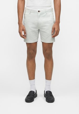 Mand iført lysegrå shorts, hvide crew-sokker og sorte slip-on sko, der står mod en ensfarvet hvid baggrund.
