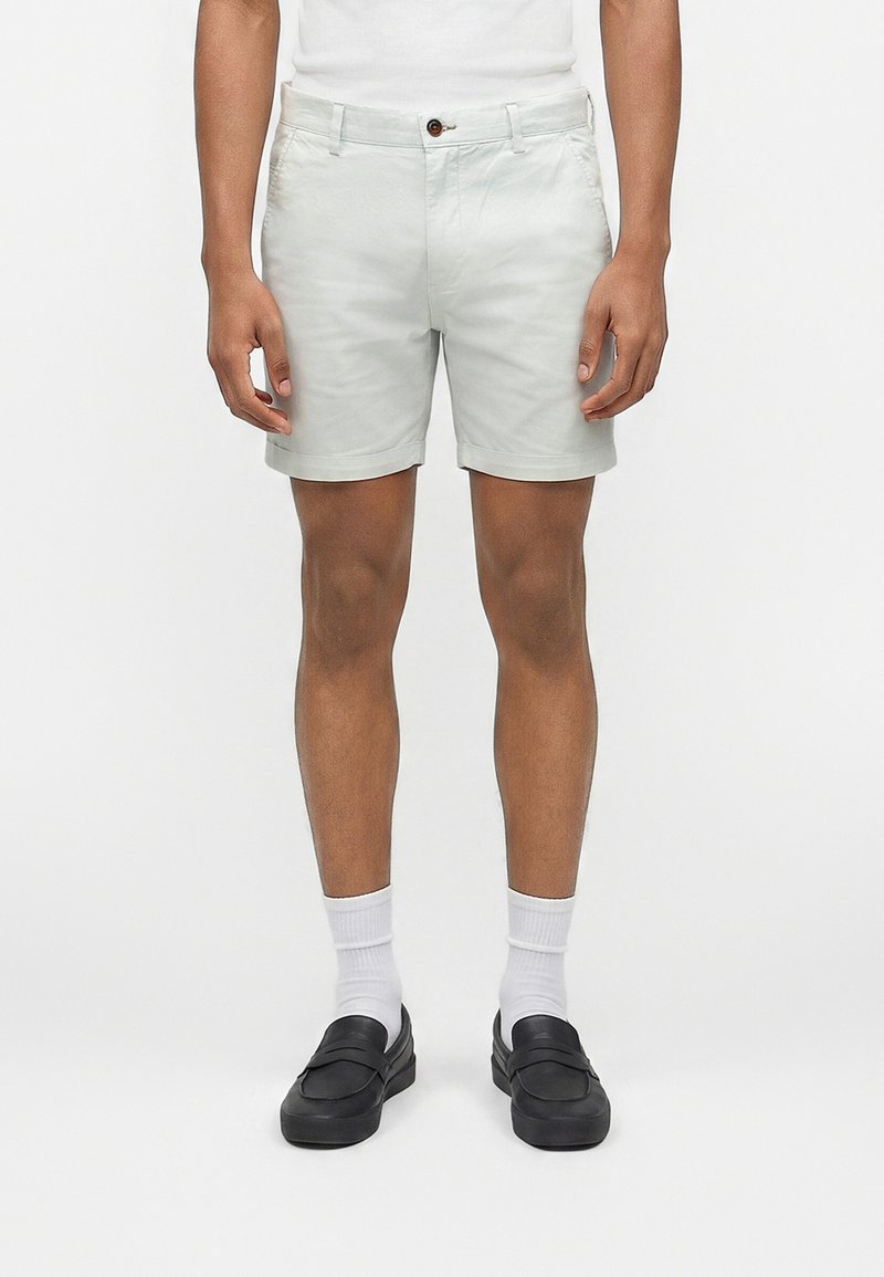 Άνδρας που φοράει ανοιχτό γκρι σορτς, λευκές κάλτσες και μαύρα παπούτσια χωρίς κορδόνια, στέκεται μπροστά σε ένα απλό λευκό φόντο.