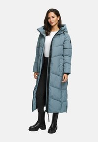 Woman wearing a long, light blue padded coat with a hood, black pants, white top, and black chunky boots standing against a plain background.