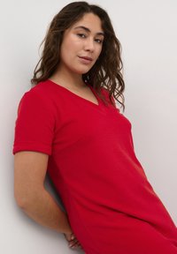 Robe courte rouge à manches courtes avec un décolleté en V et une texture douce, silhouette ajustée et manches roulées. Tissu lisse sur un fond neutre.