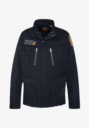 Marineblauwe jas met hoge kraag, meerdere voorkeurszakken en ritssluitingen. Kenmerkt zich door militaire patches en tekstdetails op de mouwen.