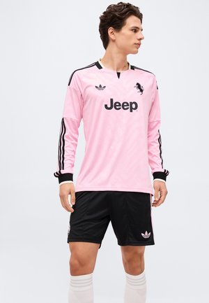 Młody mężczyzna w różowej, długorękawowej koszulce sportowej Adidas Jeep, czarnych spodenkach i białych, sięgających do kolan skarpetkach, stoi i patrzy na bok.