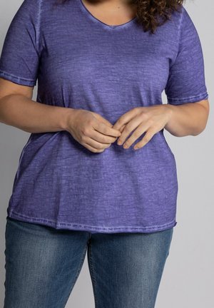 Mujer con una camiseta morada de manga corta y vaqueros azules, con las manos ligeramente entrelazadas a la altura de la cintura sobre un fondo liso.