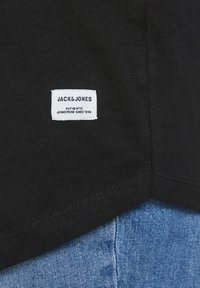 Musta kankaan T-paita, jossa on valkoinen etiketti, jossa lukee "JACK & JONES" ja "AUTENTTISTA FARKKAMERKKIÄ SINCE 1990." Näkyvät saumaristikot.