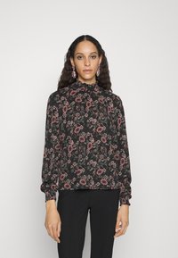Blusa floral preta, com padrões em rosa e verde, colarinho alto com franzidos e mangas longas com punhos elásticos. Combinada com calças pretas.