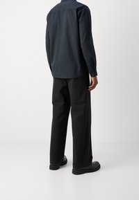 Schwarze Baumwollhose kombiniert mit einem dunklen, knopfüberdeckten Hemd. Glatte Textur, lockere Passform und gerade Beinoptik. Schwarze Schuhe runden den Look ab.