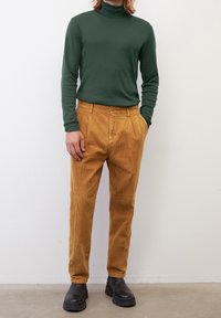 Vihreä pitkähihaisen poolopaita, joka on yhdistetty sinappikeltaisiin corduroy-housuihin. Ruskeat paksupohjaiset saappaat viimeistelevät asun. Yksinkertainen tausta.